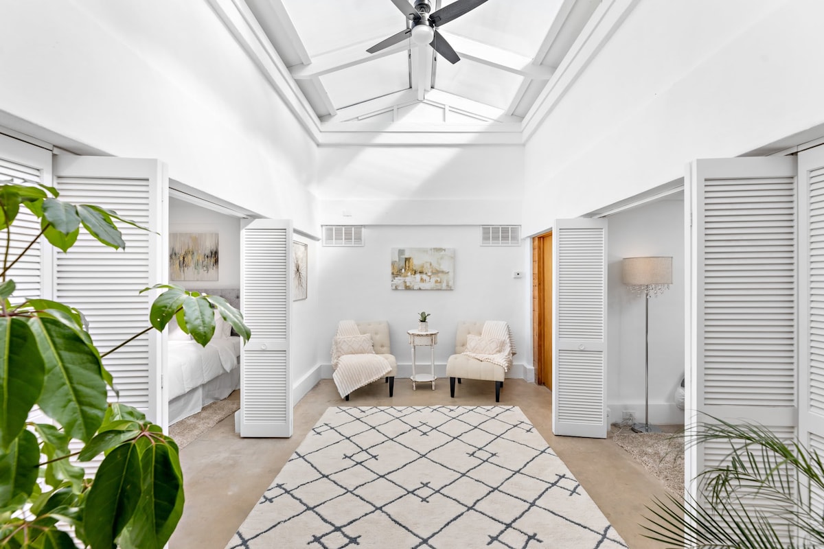 An airy entryway is illuminated by a skylight, contributing to the bright atmosphere. On either side, two spacious seating areas are arranged, featuring light-colored chairs draped with cozy throws. A patterned area rug lies on the floor, complementing the neutral decor and enhancing the welcoming environment.