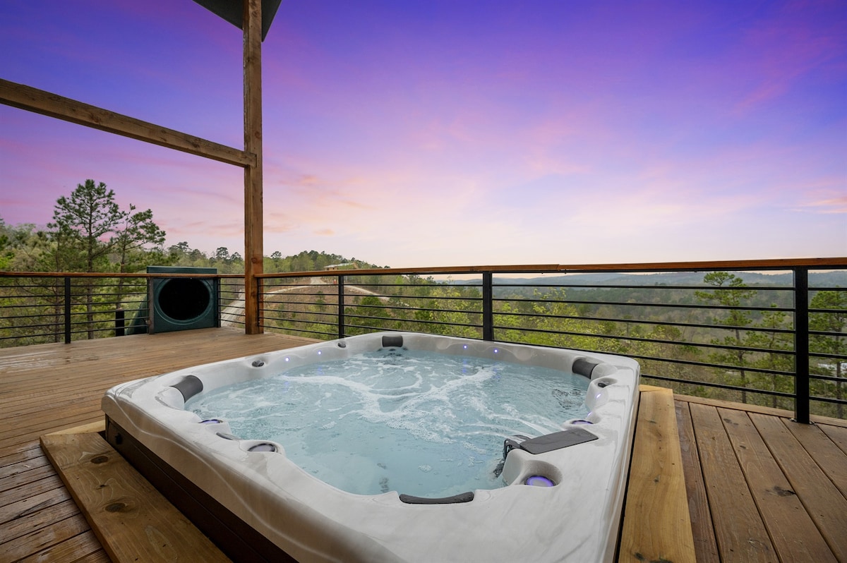 A sunken hot tub is positioned on a wooden deck, surrounded by a railing. Bubbles can be seen on the surface of the water, with trees and distant hills visible in the background under a twilight sky that transitions from soft purple to light orange.
