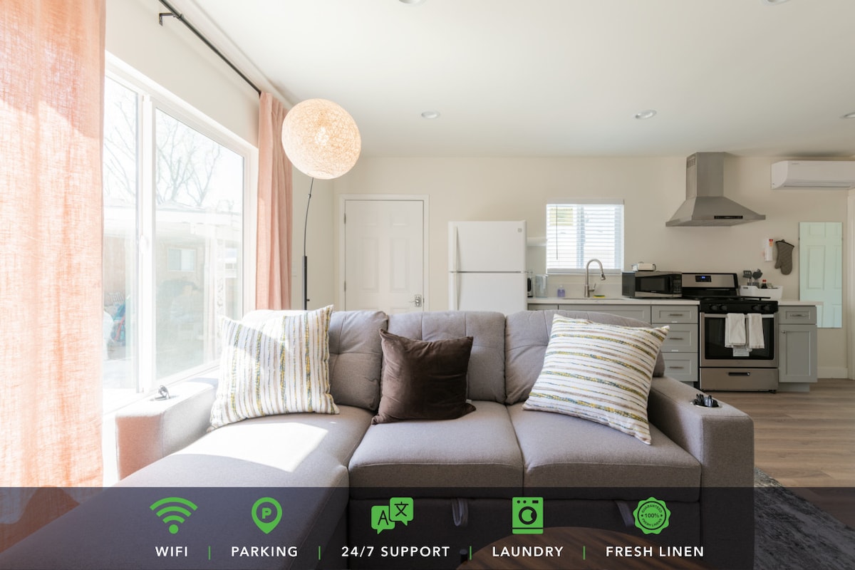A bright living area features a sectional sofa with patterned cushions, positioned near large windows allowing natural light. A modern kitchen is visible in the background with stainless steel appliances and a clean layout.
