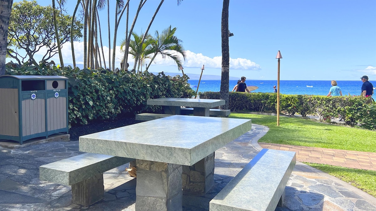 An outdoor picnic area featuring a stone table with attached benches, surrounded by tropical vegetation. The ocean is visible in the background, along with blue skies and gentle waves. A trash receptacle is positioned nearby, providing convenience for guests enjoying the coastal view.