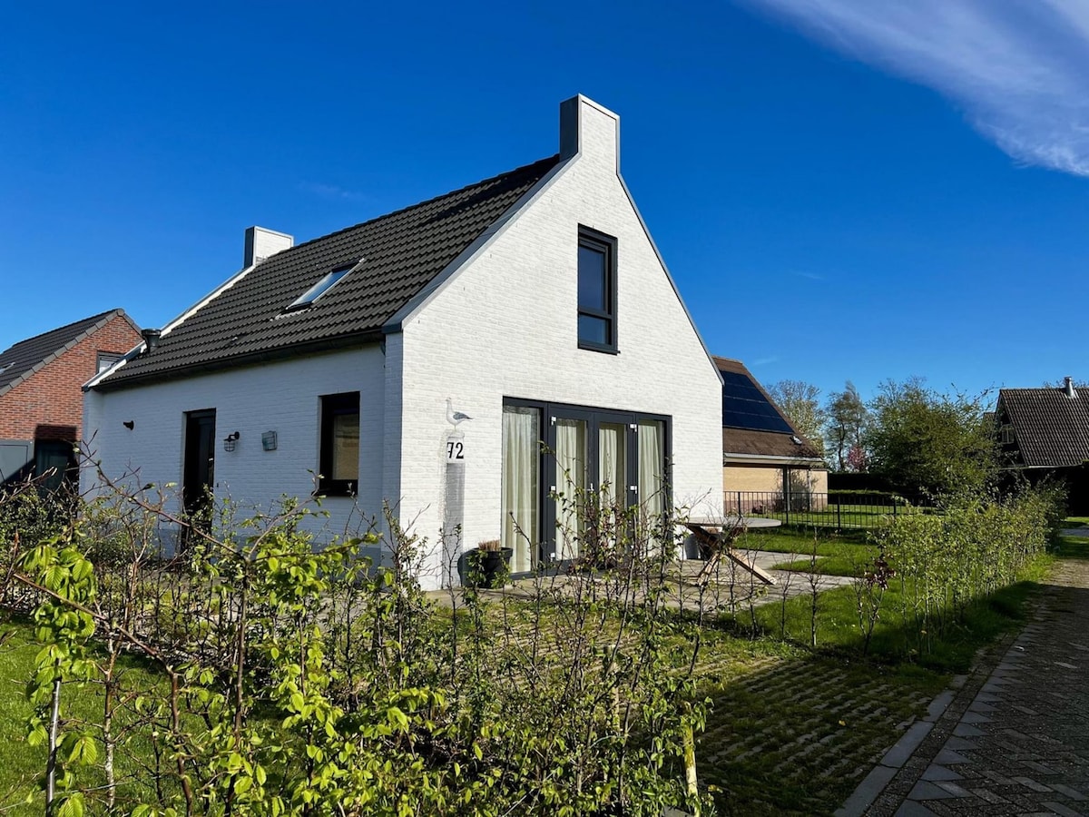 A newly constructed bungalow features a light-colored exterior with large windows, allowing ample natural light. The sloped roof adds character, while a well-maintained garden surrounds the property. The tranquil atmosphere is enhanced by the clear blue sky and surrounding greenery, inviting relaxation.