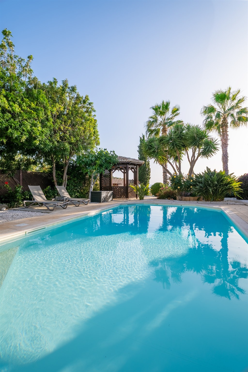 A spacious outdoor pool reflects clear blue skies, surrounded by lush greenery and tropical palm trees. Two sun loungers are positioned beside the water, while a shaded gazebo offers a relaxing retreat within the mature garden.
