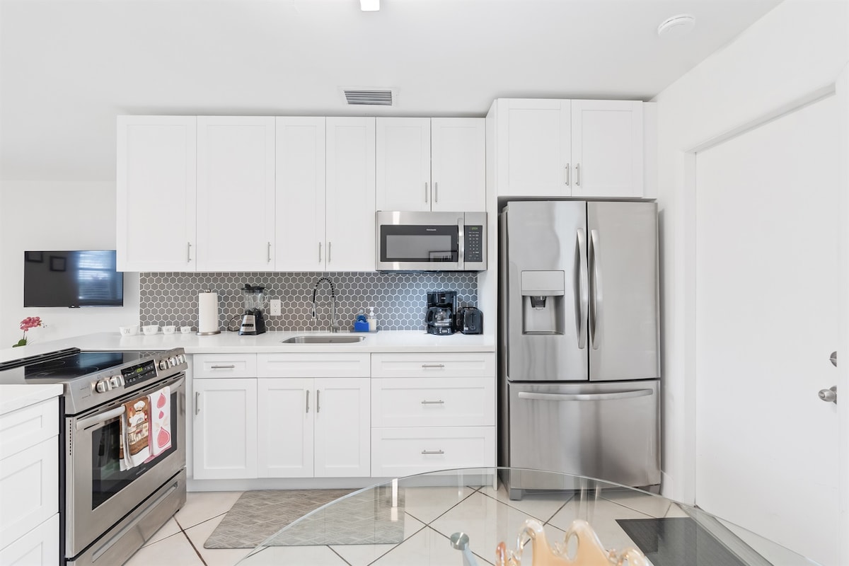 A modern kitchen is presented, featuring sleek white cabinetry and a stainless-steel refrigerator. A stove and microwave complement the space, alongside small appliances like a coffee maker. A round glass dining table is visible, with decorative items placed centrally. Neutral tile flooring completes the look.