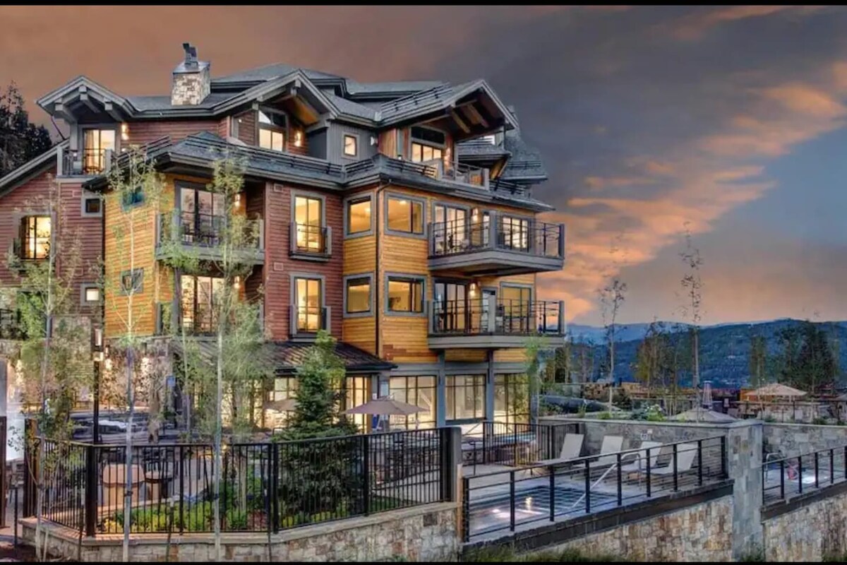The resort exterior showcases a blend of wood and stone materials, designed with multiple levels and balconies. Lush greenery surrounds the area, enhancing the inviting ambiance. A pool area is visible in the foreground, complemented by seating arrangements, with a backdrop of mountains under a colorful sunset sky.