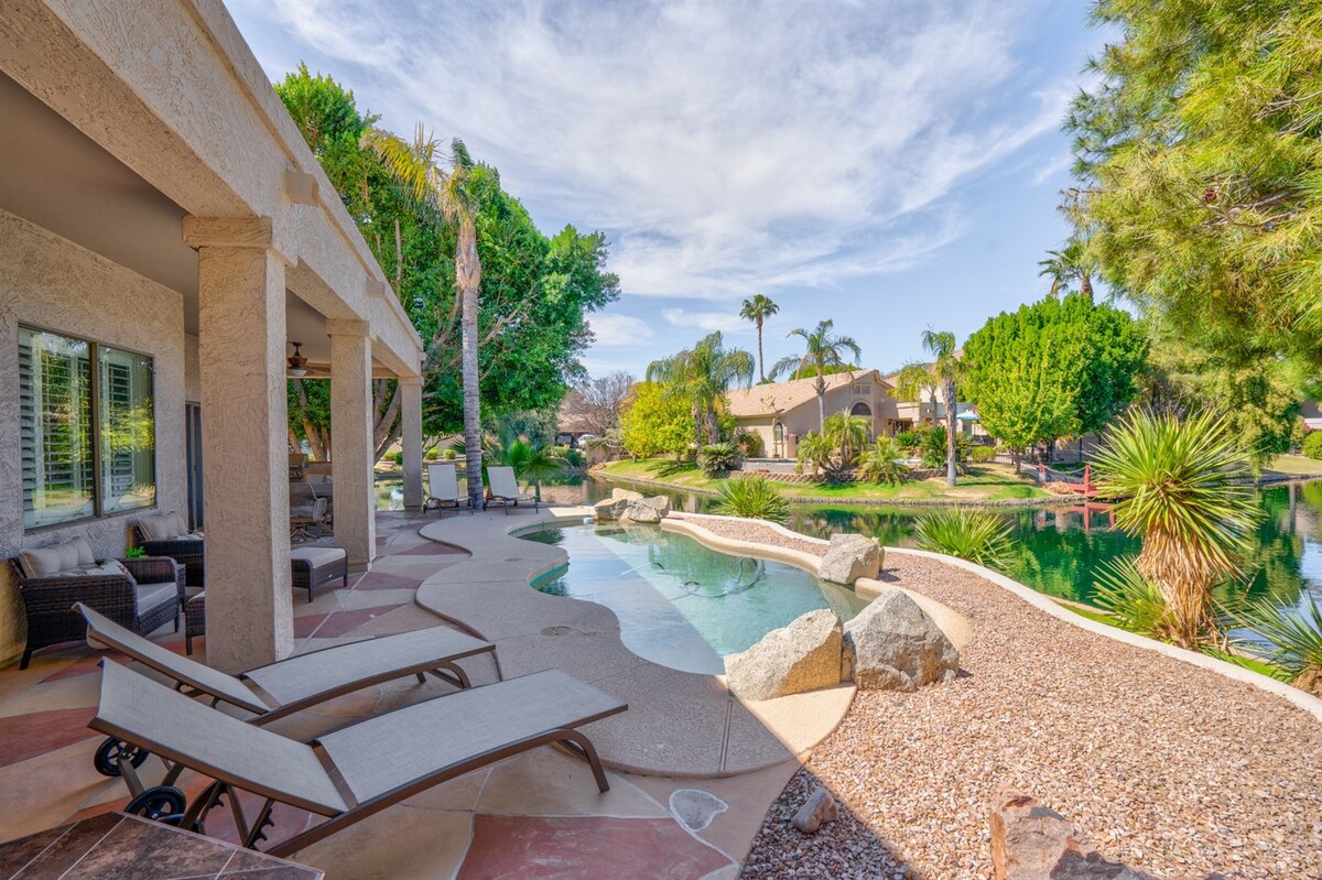 A spacious outdoor area is bordered by lush greenery, featuring a sparkling pool with a curved edge. Lounge chairs are positioned nearby, along with a clear view of the serene lake and surrounding landscape. The area is inviting for relaxation and enjoying the natural surroundings.