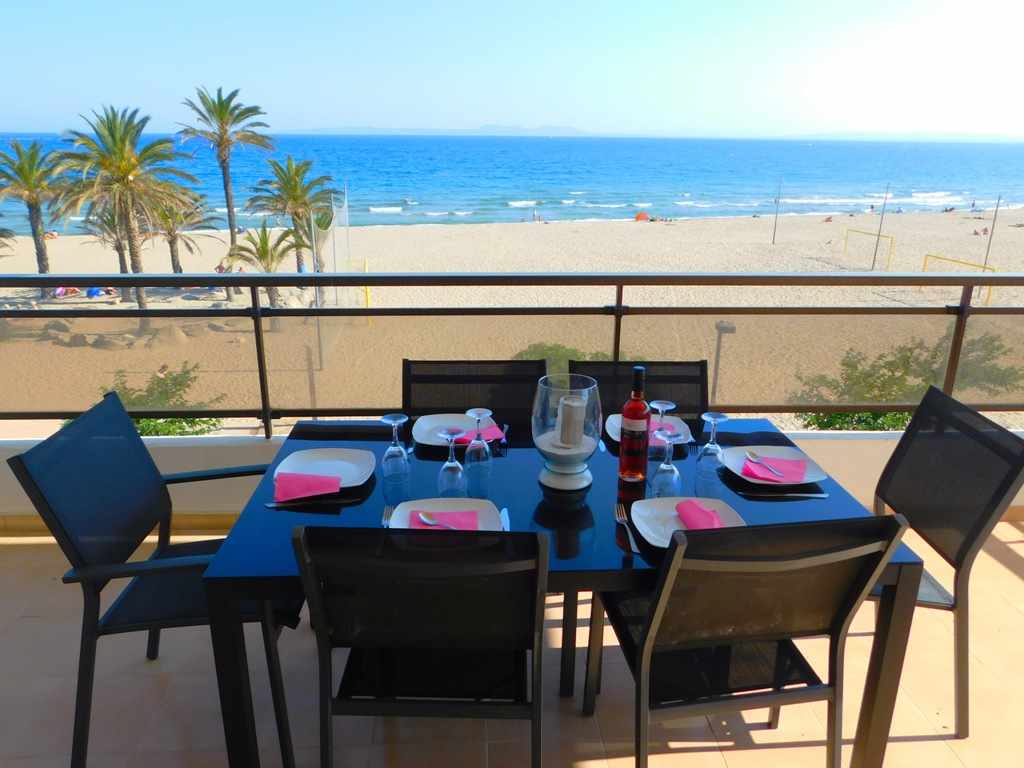 An outdoor dining area is shown with a table set for six, featuring pink napkins and glassware. A bottle of wine sits at the center. The background displays a view of the beach and palm trees, with the sea extending to the horizon.