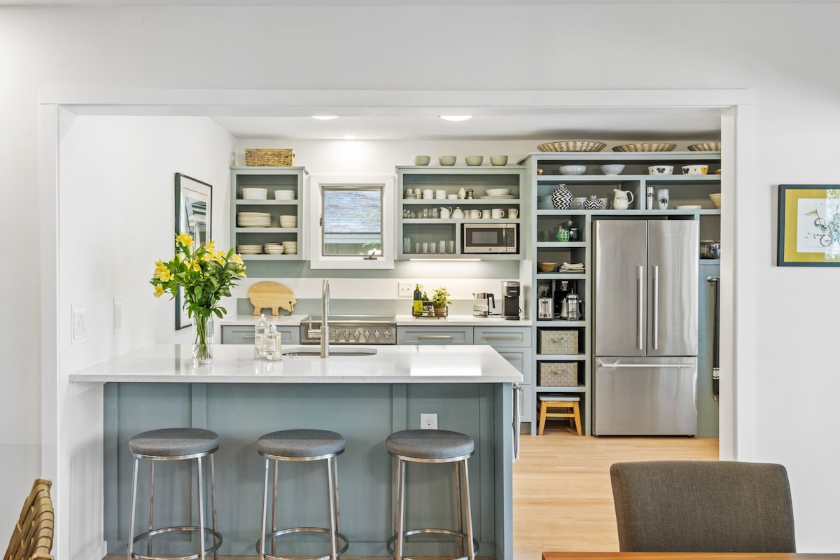 The kitchen area features modern cabinetry in soothing colors with open shelving displaying dishware. A spacious island with three bar stools offers additional seating. A stainless-steel refrigerator and clean countertop appliances complete the functional and inviting space, accented by fresh flowers in a vase.
