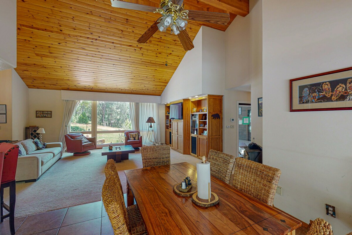 A spacious living area features high, wood-paneled ceilings, alongside large windows that flood the space with natural light. A comfortable sofa and coffee table are centered around the room, while a dining table with woven chairs provides additional seating. A warm, inviting ambiance is created by the thoughtfully arranged furnishings.