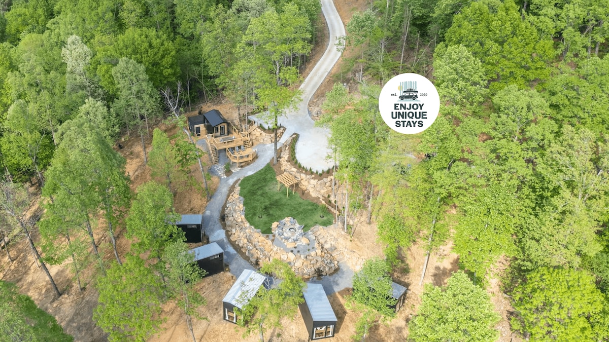 An aerial view of Treetop Escapes showcases multiple treehouse cabins nestled among lush greenery. A winding path leads to a central gathering area with a stone fire pit, surrounded by seating, while pathways connect the unique cabins in this serene, wooded setting.