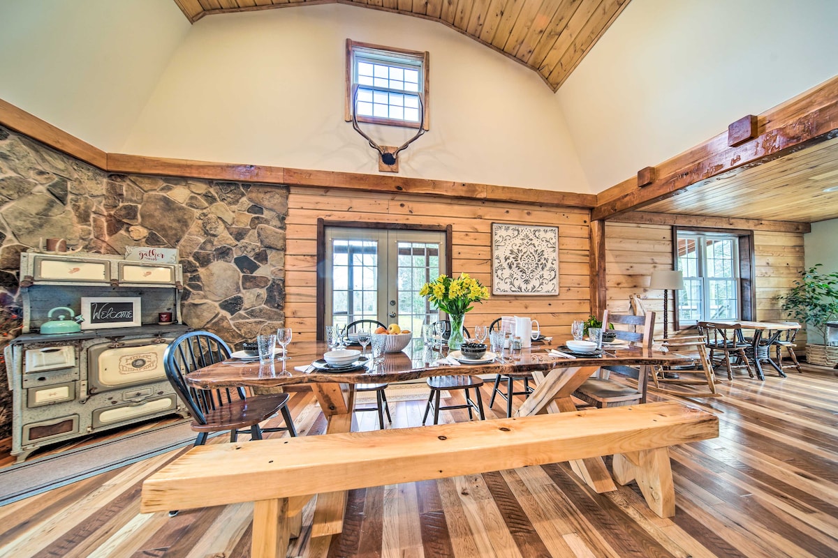 A spacious dining area features a long wooden table set for meals, surrounded by black and natural wood chairs. Sunlight filters through multiple windows, illuminating the wood-paneled walls and stone accent. A decorative piece hangs above the table, adding charm to the inviting space.