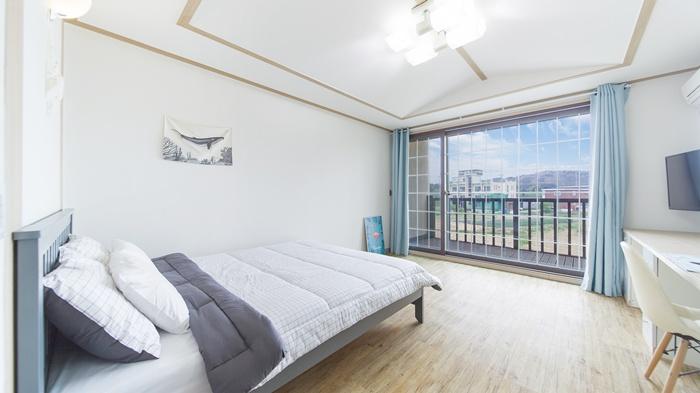 A spacious bedroom features a large window, allowing natural light to illuminate the interior. A comfortable bed with neutral bedding is positioned in the center, complemented by a simple desk and chair. Light, airy curtains frame the window, enhancing the overall brightness of the room.