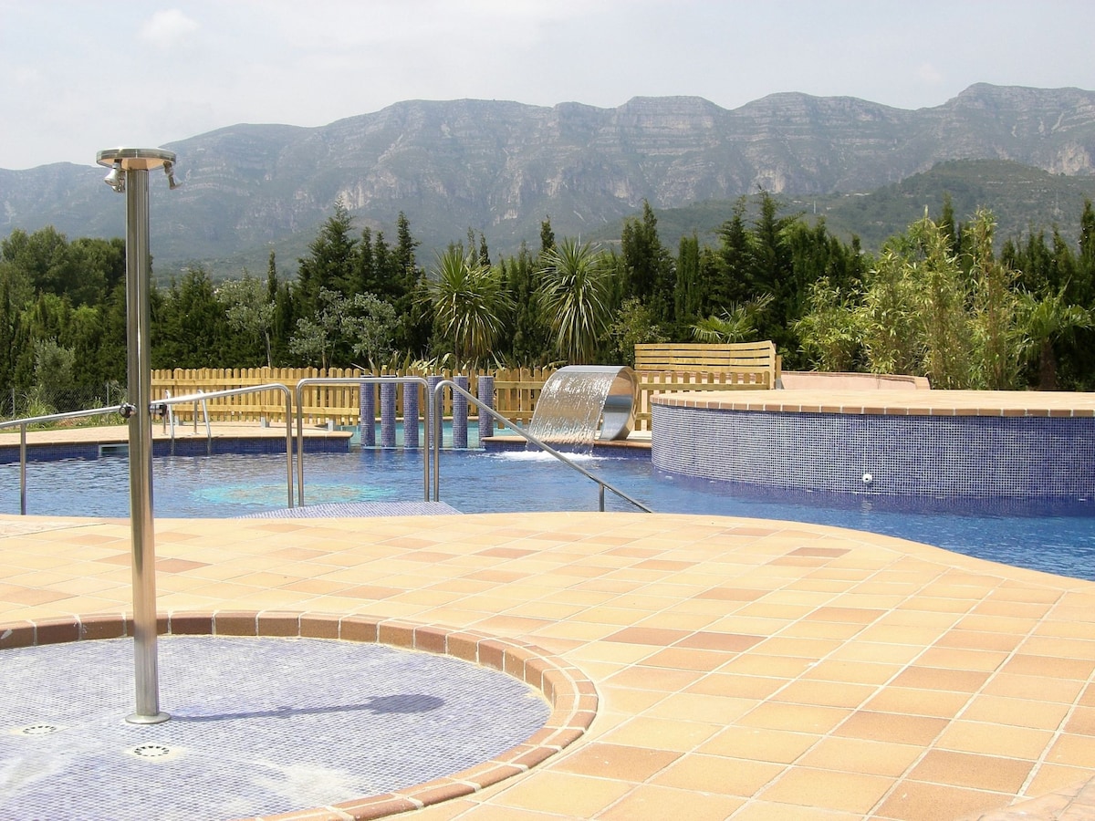 A large outdoor swimming pool with a gently sloping entrance features a waterfall and shallow circular area. Surrounding greenery and mountains provide a natural backdrop. A wooden bench is visible on the pool deck, offering a space for relaxation.