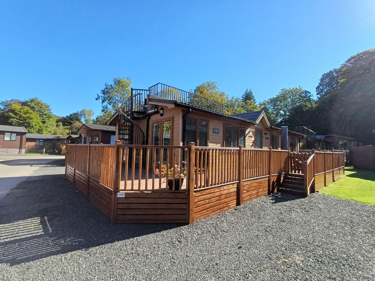 A wooden lodge is showcased with a spacious, enclosed decking area. The roof terrace is visible, accessed by spiral steps. Surrounding greenery enhances the outdoor setting, while clear blue skies provide a bright atmosphere.
