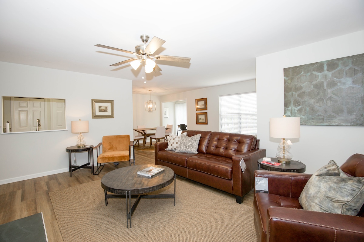A spacious living area features a ceiling fan and comfortable seating options, including two sofas and an armchair. A round coffee table sits at the center, with natural light filtering through nearby windows. A dining area is visible in the background.