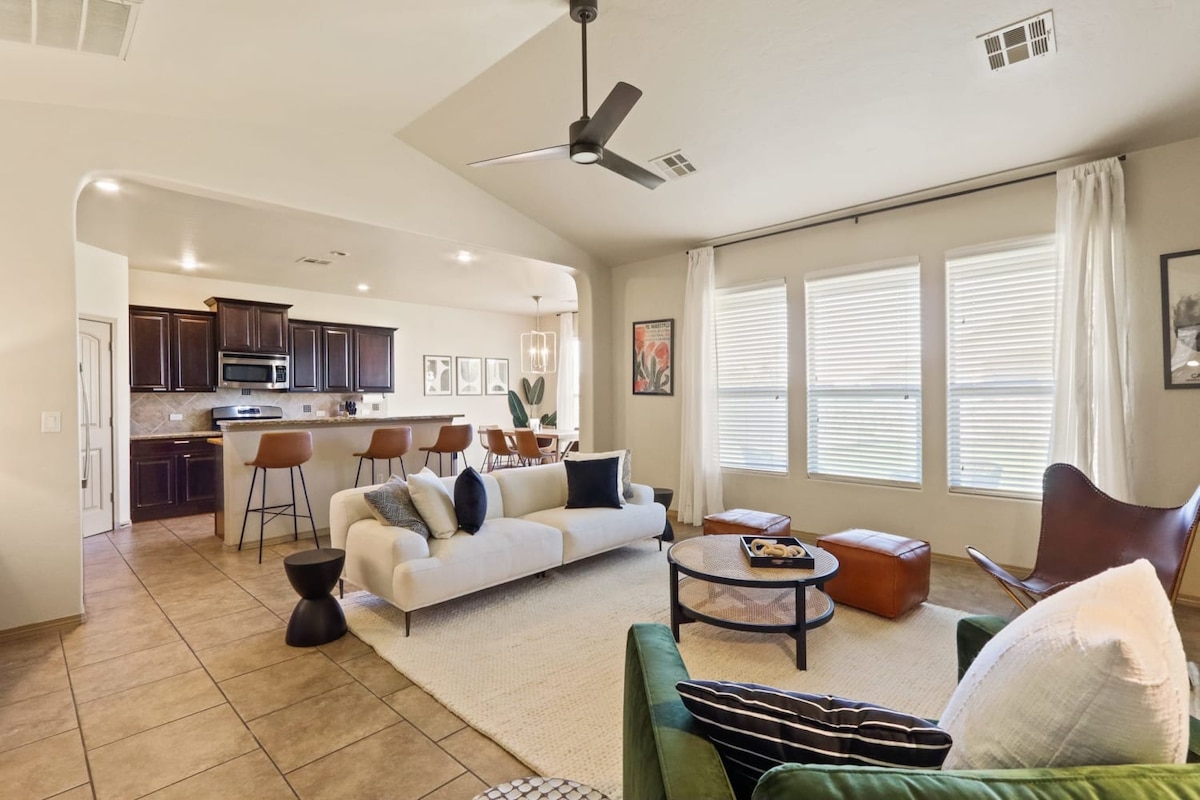 The spacious living area is adorned with natural light from large windows, featuring a comfortable sofa and accent chairs. A stylish coffee table occupies the center, while the adjacent dining area and kitchen are visible, showcasing modern furnishings and elegant design.