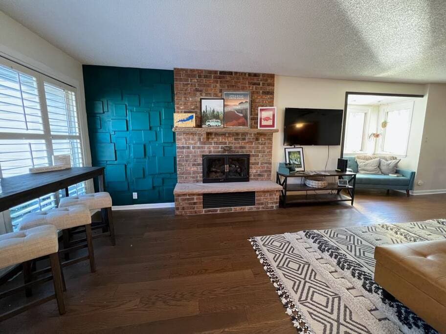 A warm, inviting living area features a textured teal wall and a brick fireplace, creating a cozy focal point. A light hardwood floor complements the space. A dining table with high-backed stools and a comfortable seating area is visible, with natural light streaming in from multiple windows.