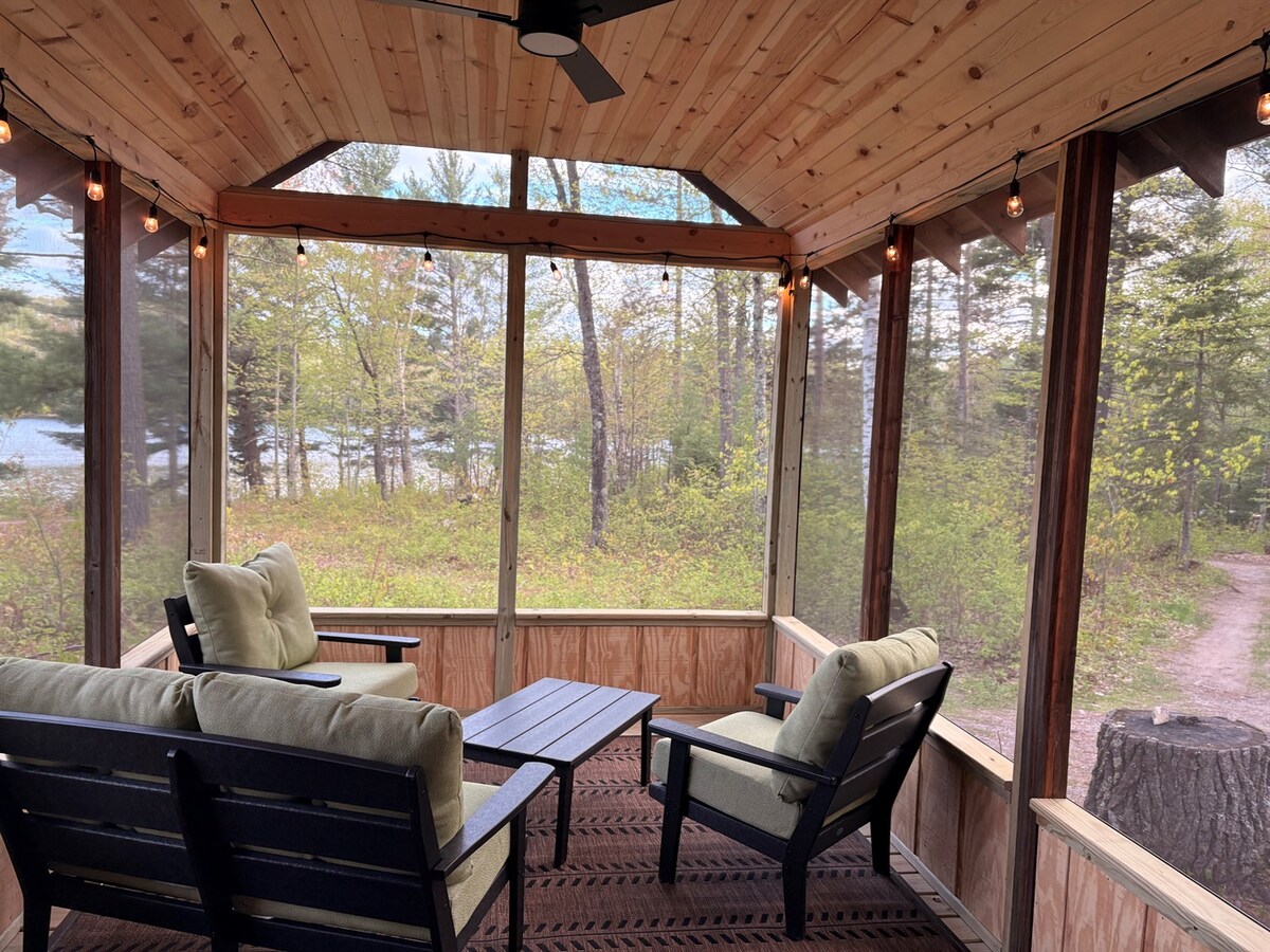 A small screened porch features comfortable seating with soft cushions and a dark rug. Natural light filters through the screens, revealing lush greenery outside. The ceiling is adorned with wooden paneling and a ceiling fan, enhancing the inviting atmosphere.