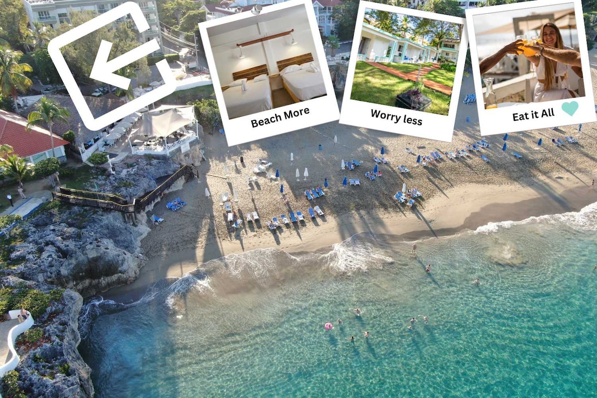 An aerial view showcases a sandy beach lined with sun loungers, gently touched by serene waves. Colorful umbrellas provide shade for visitors enjoying the shoreline. The clear blue water reflects sunlight, inviting a sense of relaxation and adventure.