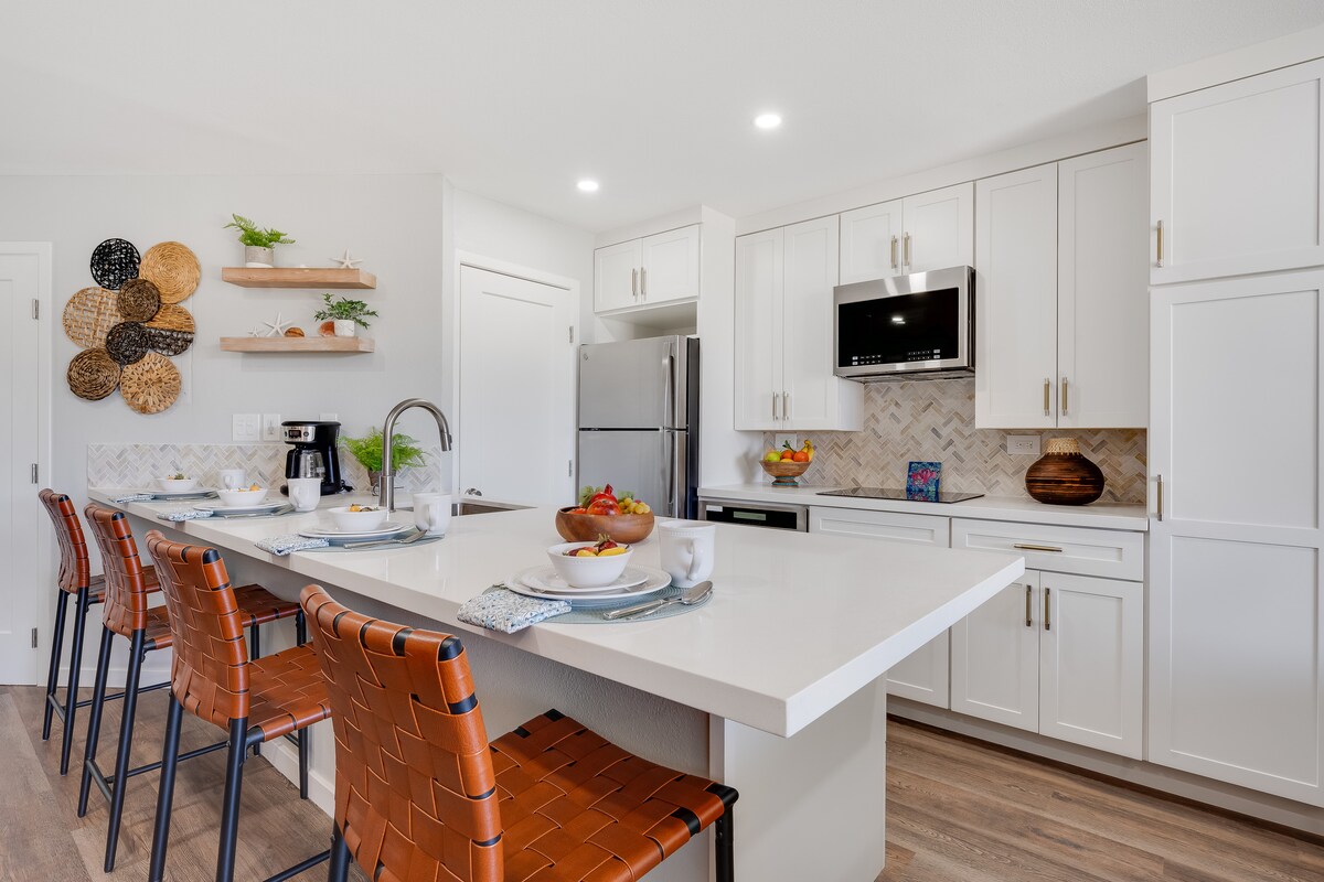 A modern kitchen features white cabinetry with polished hardware and a large central island. Four high-backed, brown leather bar stools are positioned around the island. The countertop showcases a bowl of fresh fruit and elegant place settings, while stainless steel appliances and stylish backsplash tiles complete the space.