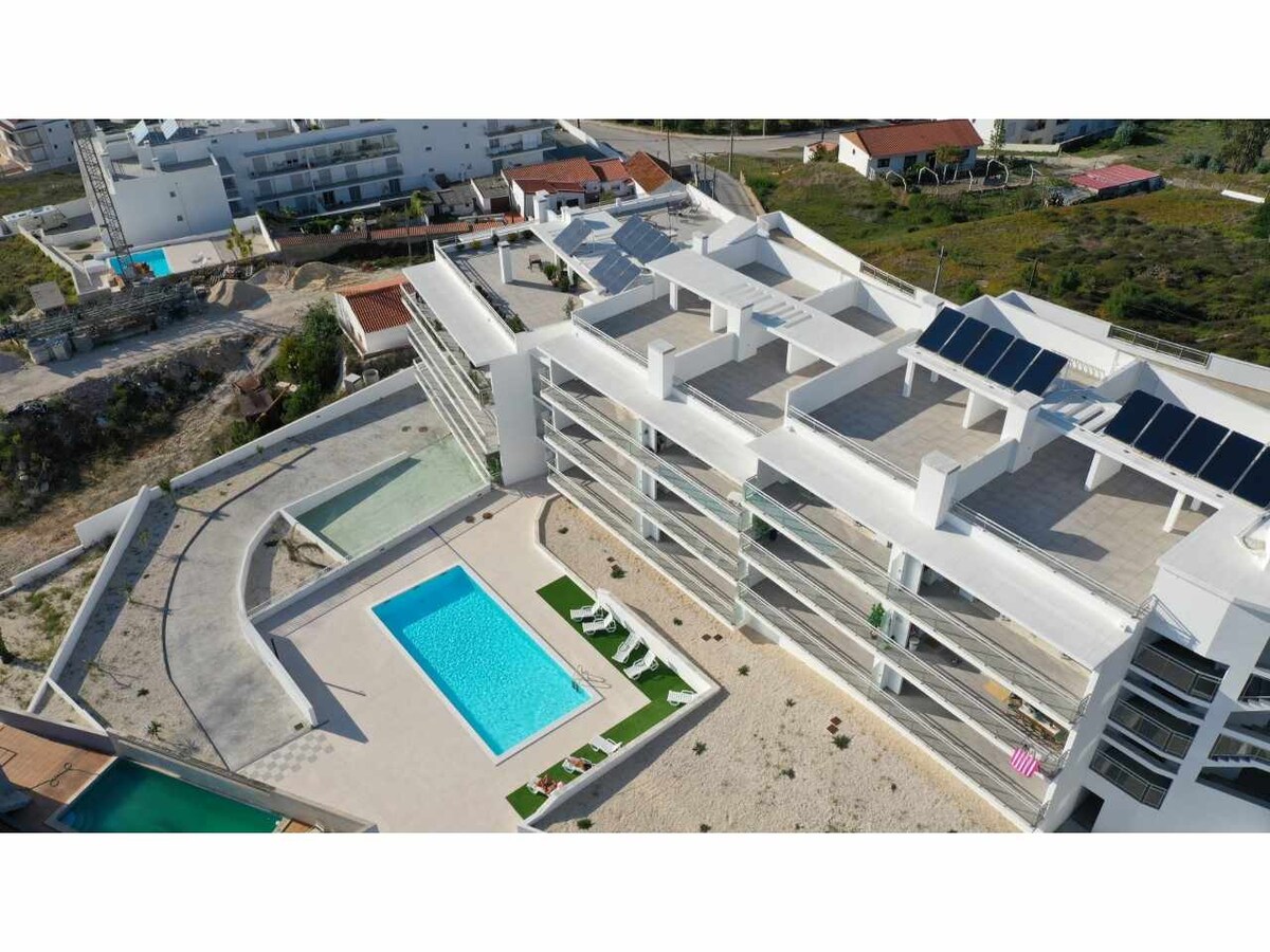An aerial view of a modern apartment complex features multiple terraces and a swimming pool area, surrounded by landscaped gardens. The clear blue pool area is complemented by sun loungers. Solar panels are visible on the rooftops, highlighting sustainable architecture.