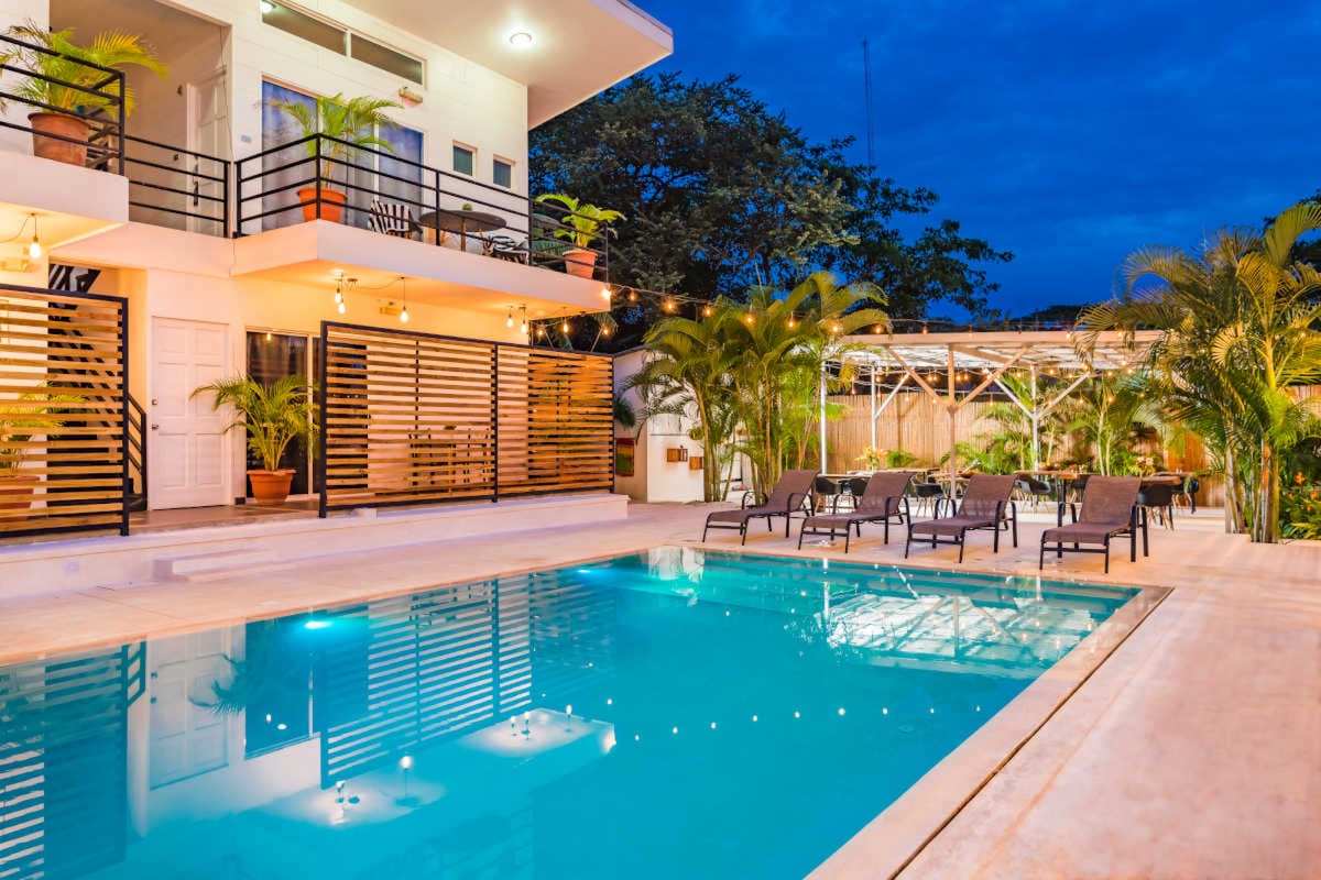 A well-lit pool area is surrounded by lounge chairs and tropical plants. The modern building features a second-floor balcony with sliding doors, while string lights create a cozy atmosphere. The clear water of the pool reflects the evening sky and surrounding greenery.