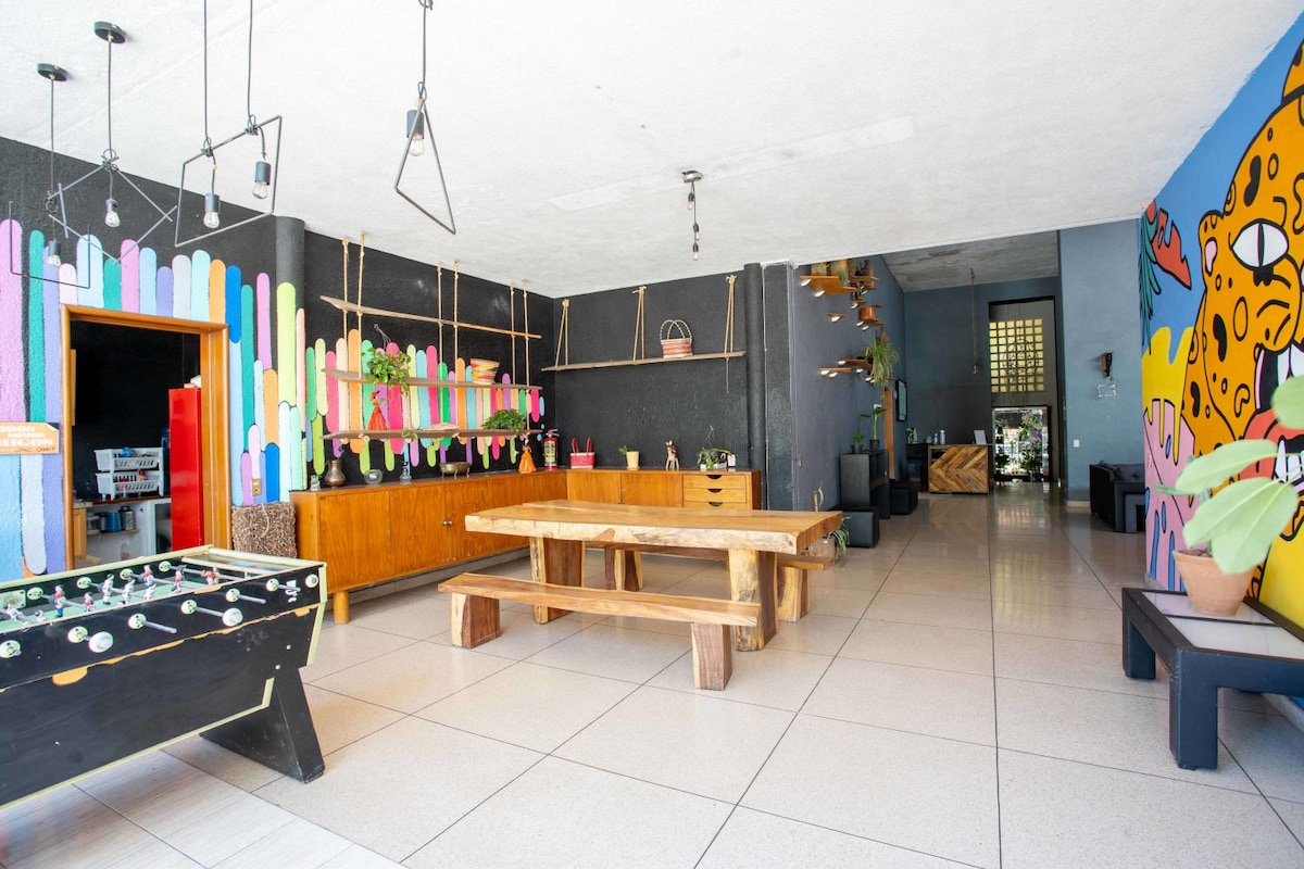 The communal area features colorful murals against dark walls, creating an energetic atmosphere. A large wooden table is centrally positioned for gatherings, while foosball and seating areas are accessible. Ambient light filters through large windows, enhancing the spacious layout of the room.