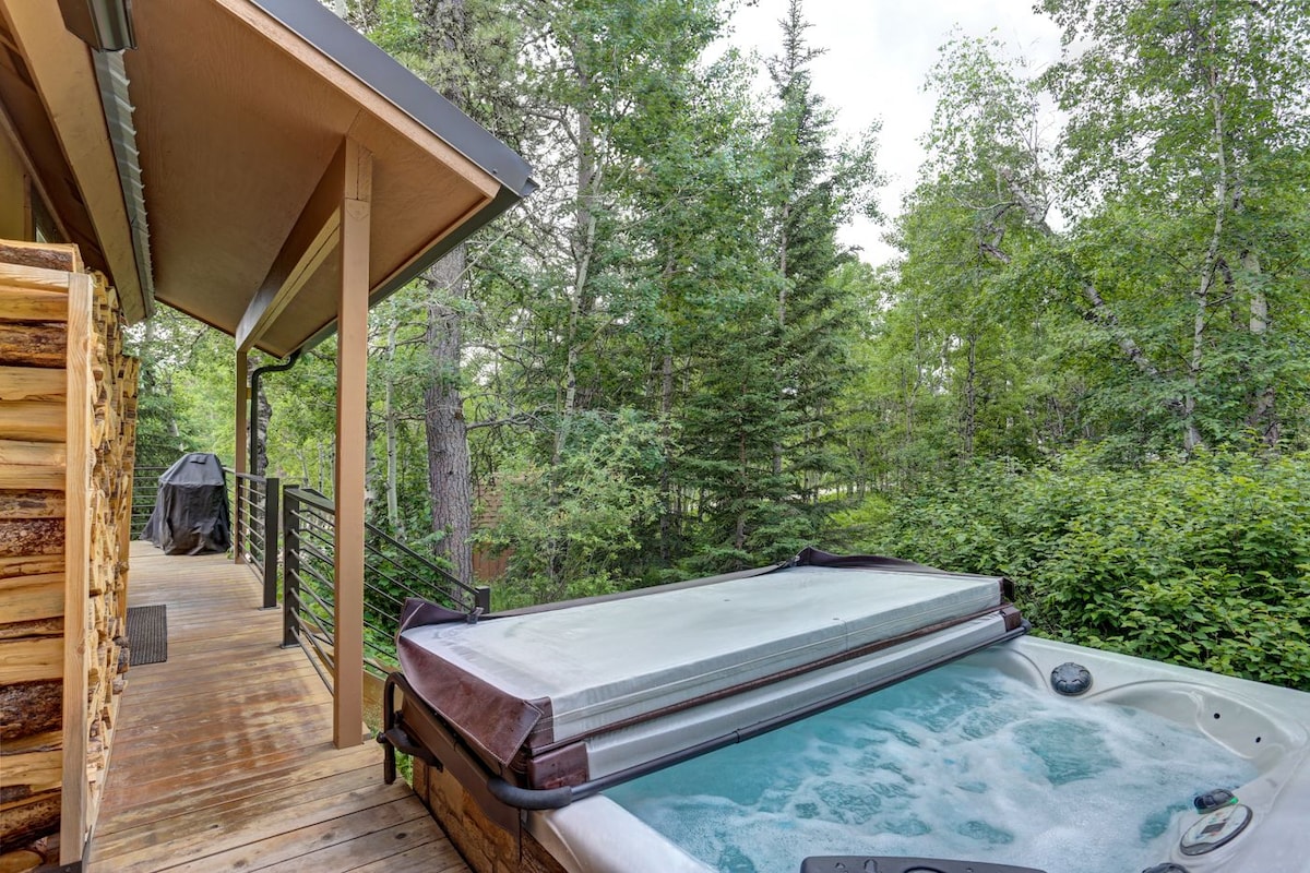 An outdoor hot tub is situated on a wooden deck, surrounded by lush green trees and foliage. The cabin is partially visible in the background, with a portion of a covered grill area also included.