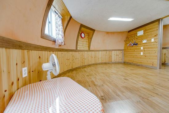 A uniquely shaped interior space features gently curved walls adorned with warm wooden paneling. A small fan is positioned near a checkered cushioned seating area. Natural light enters through a window with floral curtains, enhancing the serene environment.