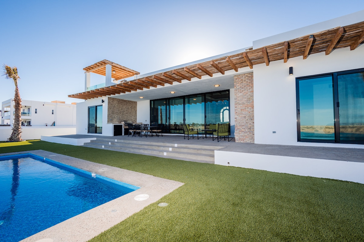 A modern home exterior is highlighted by a spacious patio and large glass doors that invite natural light. A private pool is visible in the foreground, with manicured green grass surrounding the area. A shaded seating arrangement offers a view of the pool and outdoor surroundings.