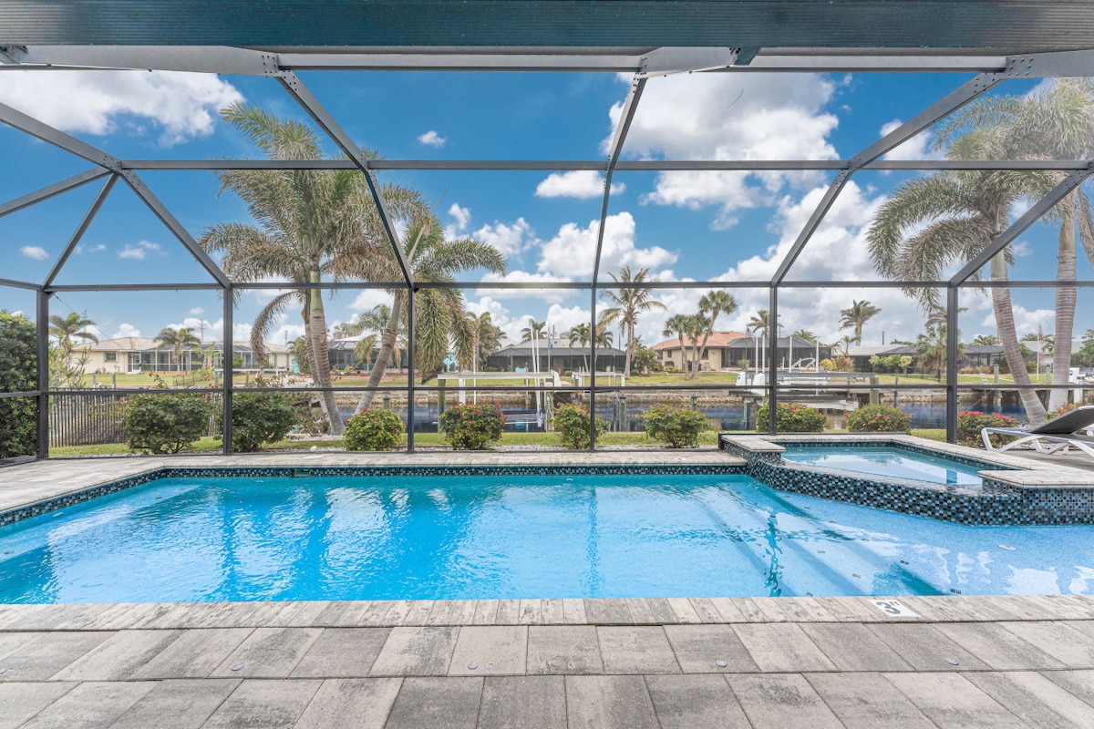 The outdoor area features a heated saltwater pool and spa, enclosed within a screened lanai. Lush greenery and palm trees surround the pool, creating a serene environment with views of the canal waterfront and clear blue skies visible through the spacious screens.