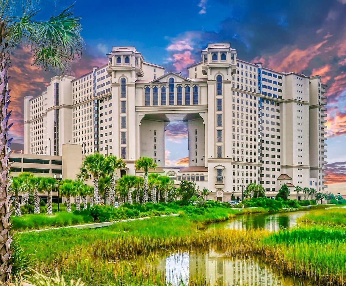 The expansive resort building features a unique architectural design with arched entrances, set against a vibrant sunset sky. Lush greenery and palm trees surround the property, while a tranquil waterway reflects the structure, emphasizing its grandeur and serene surroundings.