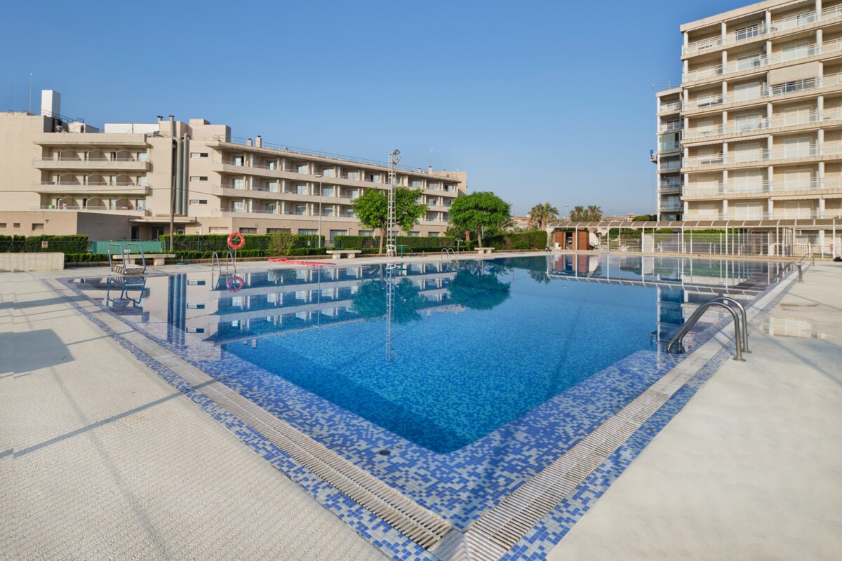 A large swimming pool is depicted, featuring clear blue water reflecting the sky. Surrounding the pool area are well-maintained gardens and residential buildings. Loungers are positioned nearby, providing spaces for relaxation under the open sky.
