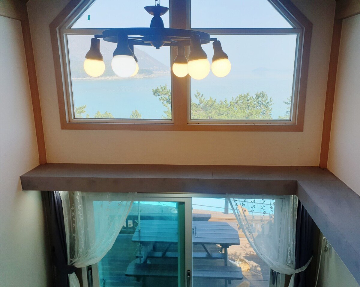 A well-lit interior features a modern chandelier hanging from a sloped ceiling. Large windows provide a stunning view of the sea, while sheer curtains frame the glass doors leading to an outdoor seating area. Natural light fills the space.