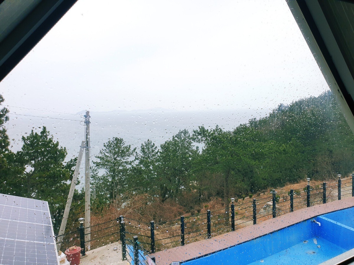 A view of the sea is captured through a window, with raindrops visible on the glass. Green trees are seen in the foreground, while the serene water stretches towards the horizon. A section of a swimming pool is displayed in the lower corner.