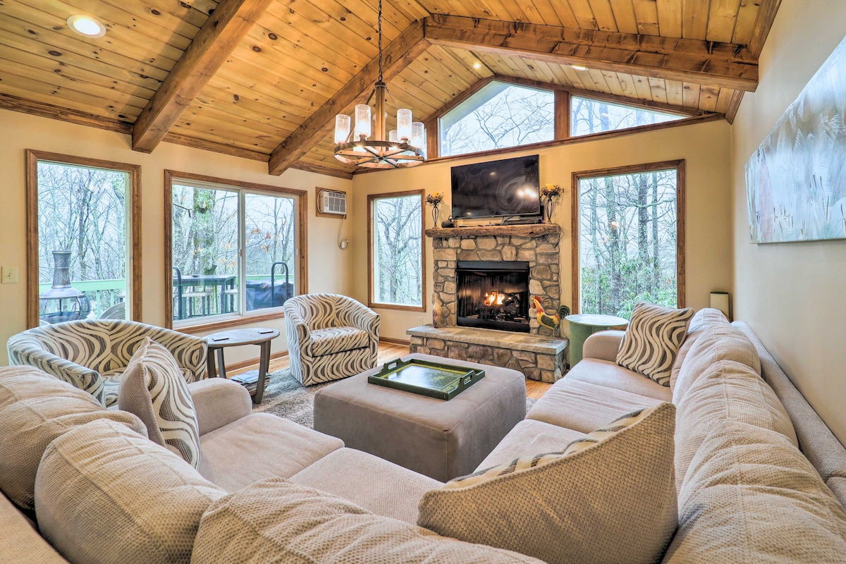 A cozy living area features a spacious sectional sofa and two accent chairs, oriented towards a stone fireplace. Large windows offer views of the surrounding trees, while a rustic chandelier hangs from the wooden ceiling, contributing to the room's inviting ambiance.