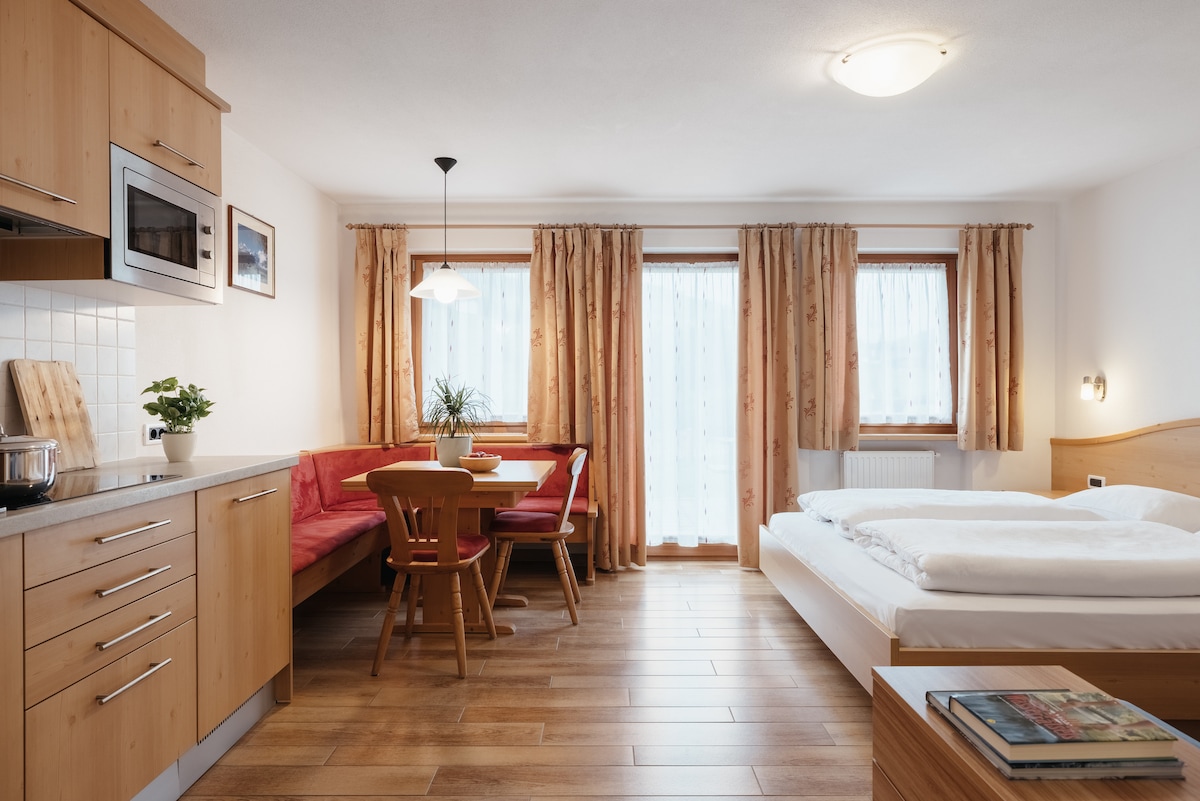 The studio features a light wood kitchen area with a built-in microwave and dishwasher. A dining nook with red upholstery complements a small table and wooden chairs. A spacious bed is positioned next to large windows adorned with light curtains, allowing natural light to enter the room.