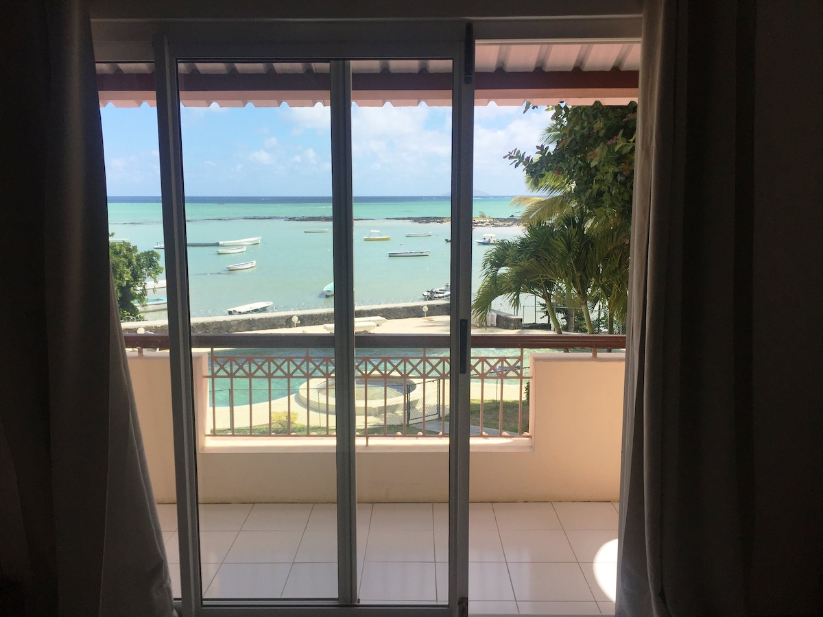A glass sliding door opens to a private terrace with a view of the tranquil turquoise lagoon. Calm waters are dotted with small boats, surrounded by lush greenery. Bright sunlight filters through the open doorway, enhancing the serene coastal scene.