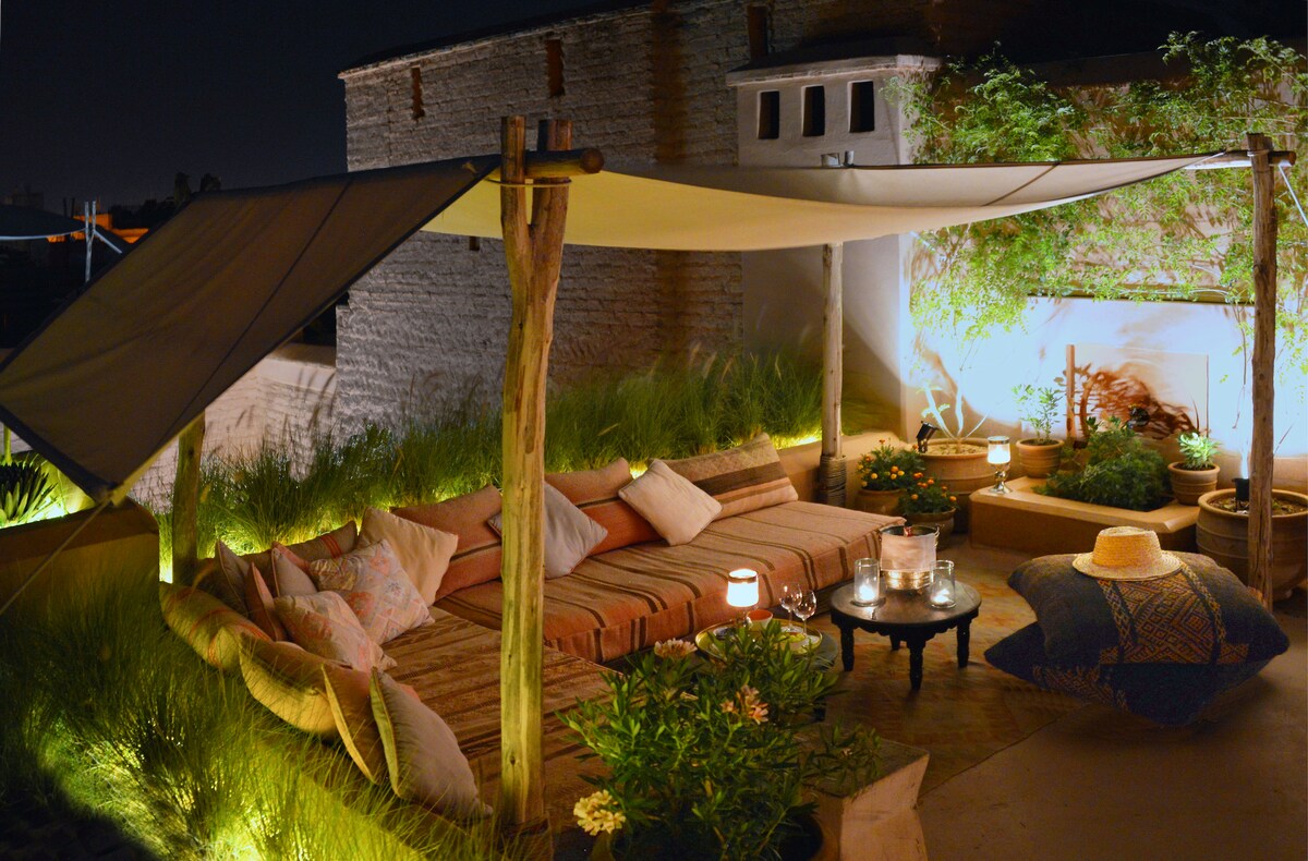 An intimate outdoor seating area is illuminated by warm lighting, featuring low cushioned seating arranged around a dark table. Potted plants and greenery create a serene environment, complemented by decorative accents. A canopy provides shade, enhancing the cozy atmosphere for evening relaxation.