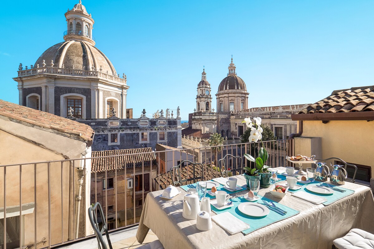 A spacious terrace is furnished with a dining table set for a meal, featuring dishes and utensils arranged neatly. The view captures historic domes and rooftops under a clear blue sky, providing a sense of connection to the vibrant surroundings.