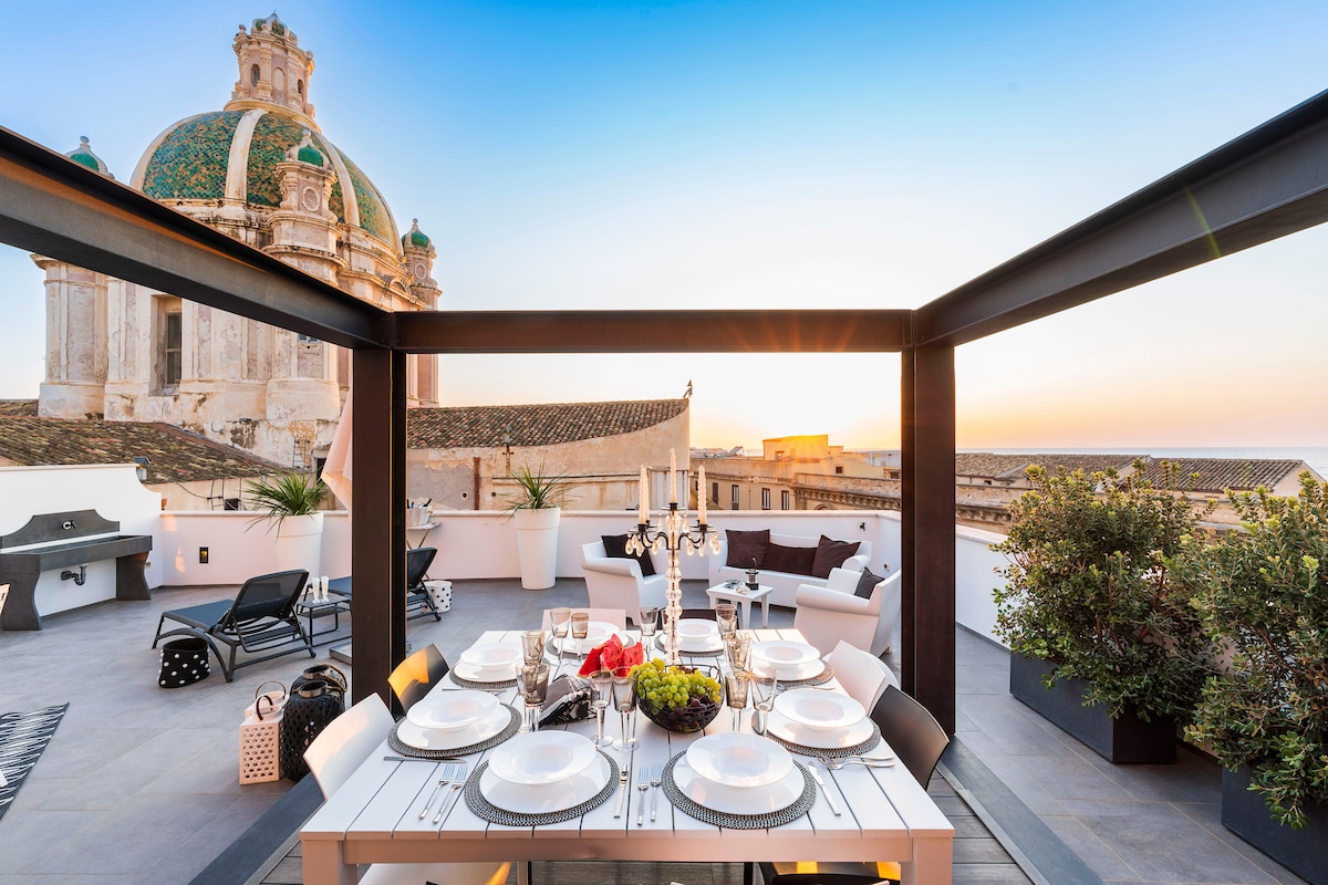 An elegant terrace features a dining table set for al fresco meals, surrounded by stylish seating and planters. The stunning dome of a nearby building is visible in the background, while a sunset casts warm light over the area.