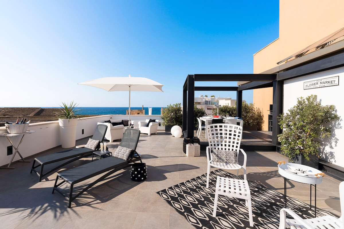 A spacious terrace is presented, featuring sun loungers and a covered seating area. An umbrella provides shade, while plants add greenery. A grill is positioned near a dining table, with stunning views of the sea visible in the background under a clear blue sky.