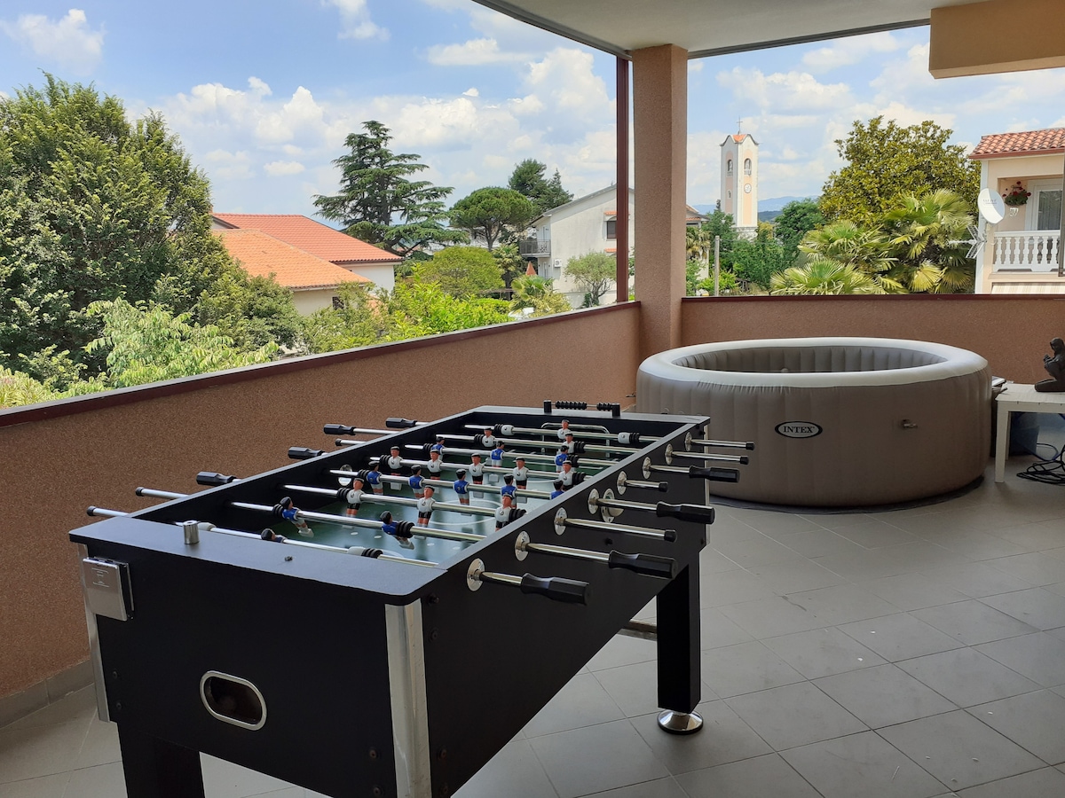 A spacious terrace features a foosball table beside a circular hot tub. Lush greenery outlines the view, with a distant clock tower visible. The area is designed for relaxation and leisure, with ample natural light filtering through the surrounding space.