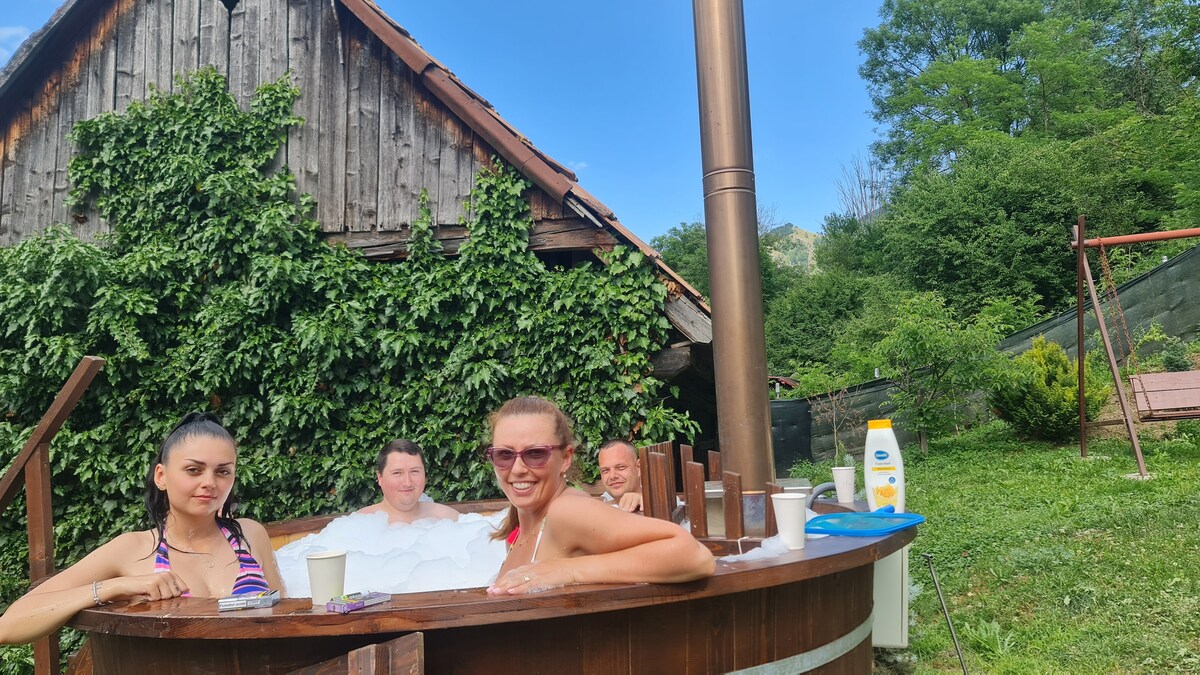 An outdoor hot tub is surrounded by lush greenery and a rustic wooden structure in the background. Passengers are seen enjoying the warm water, with a chimney positioned nearby. Soft sunlight illuminates the scene, creating a relaxing environment for leisure.