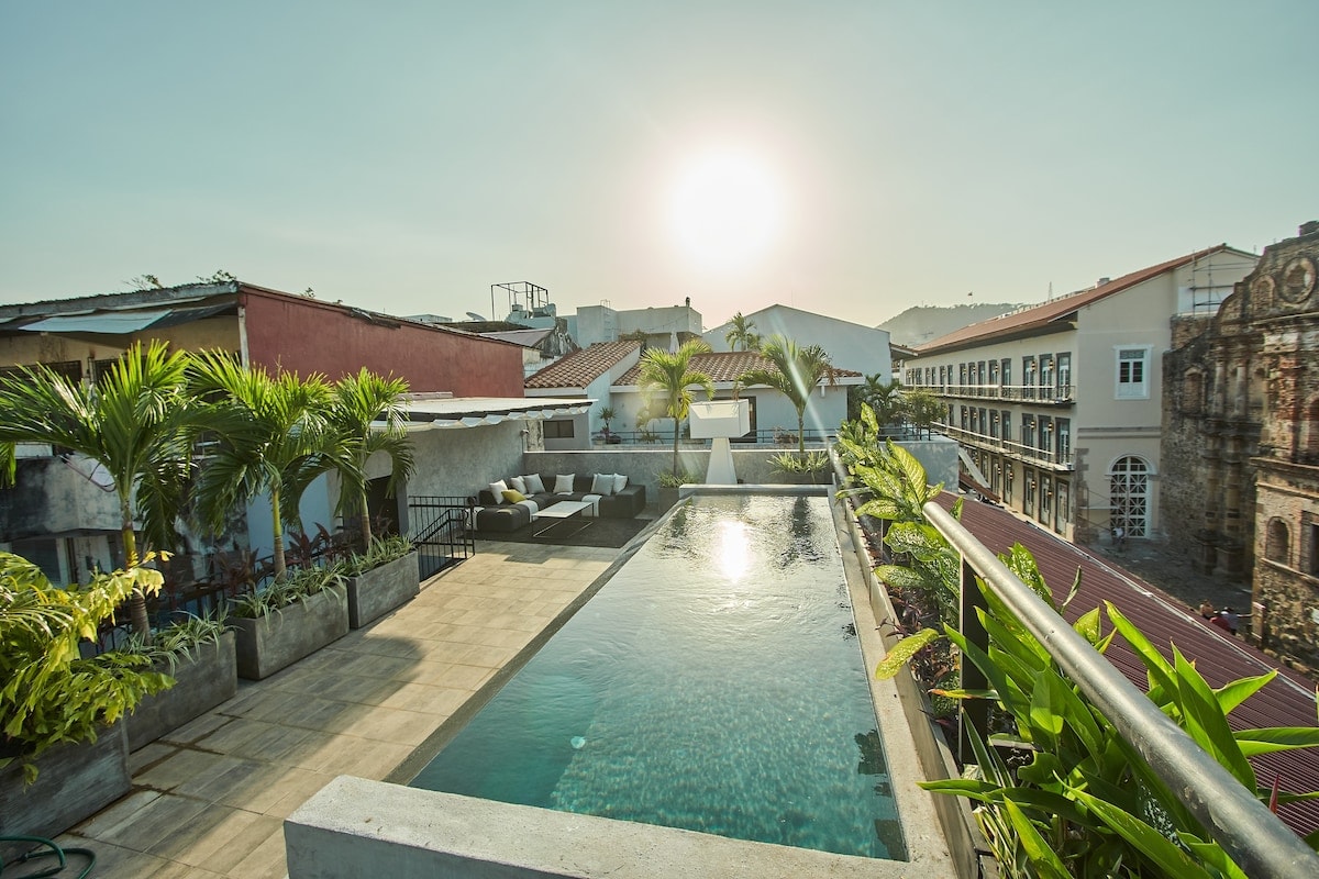 An infinity pool is showcased on the rooftop, surrounded by lush greenery and comfortable outdoor seating. The sun sets over the horizon, casting a warm glow on the serene landscape. The view includes neighboring buildings and the vibrant atmosphere of Casco Viejo.