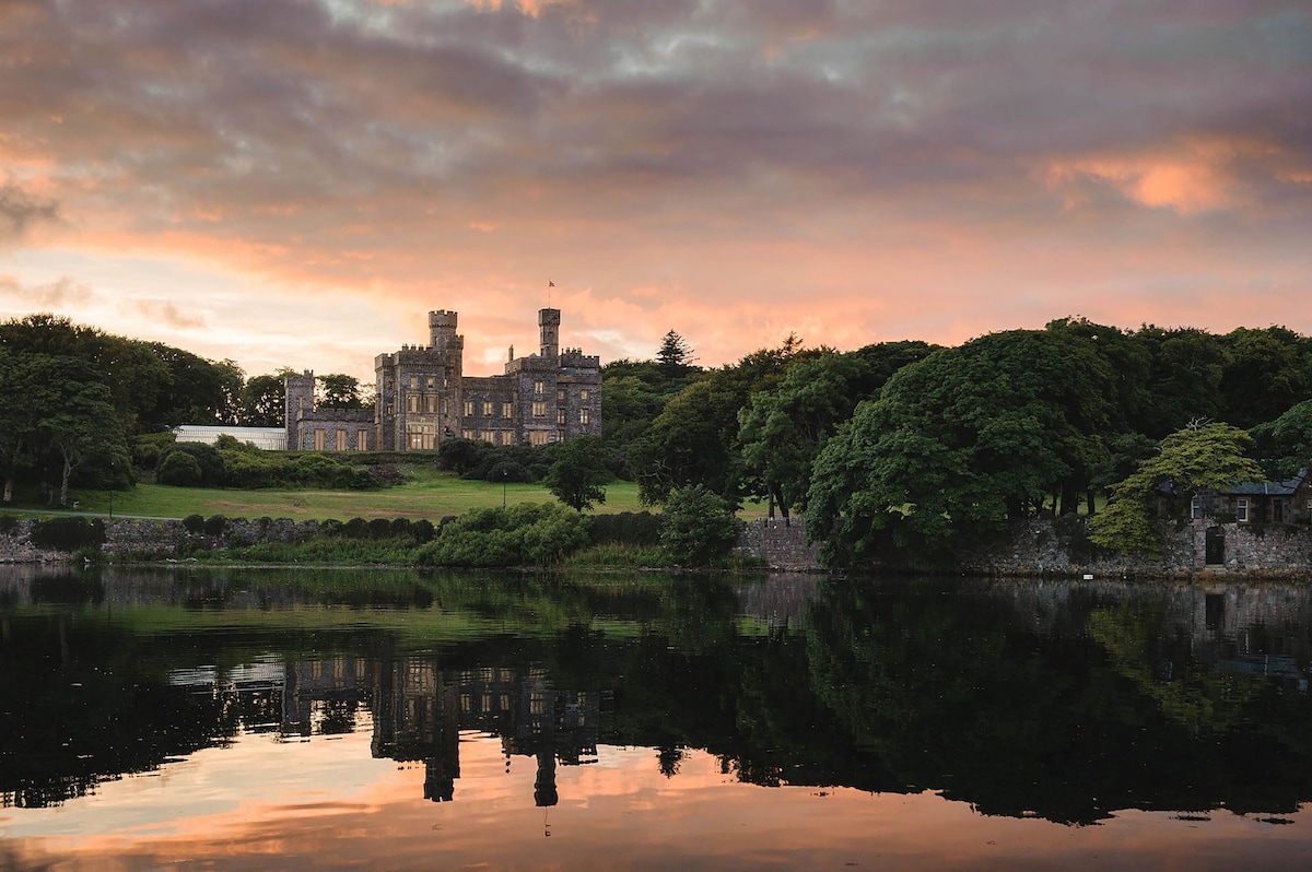 The grand structure of Lews Castle is reflected on the calm surface of the adjacent water. Surrounding lush greenery and trees frame the scene, with a colorful sunset sky adding warmth to the backdrop.