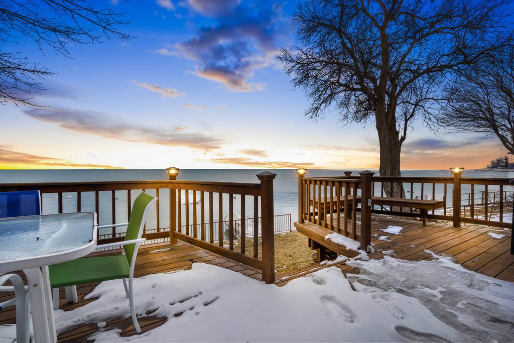 A wooden deck is shown overlooking a calm body of water with a vibrant sunset in the background. Snow is present on the ground, and outdoor seating is visible, including a table and a green chair. Warm light is emitted from lanterns along the railing.