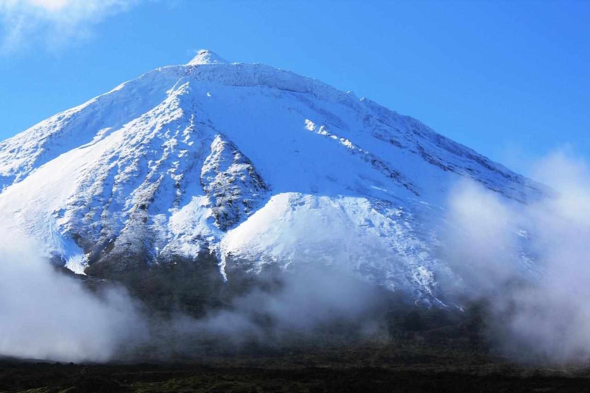 A snow-covered mountain peak towers against a clear blue sky, with wisps of white clouds drifting near its base. The slopes exhibit a contrast of rock and ice, reflecting the natural beauty of the landscape.