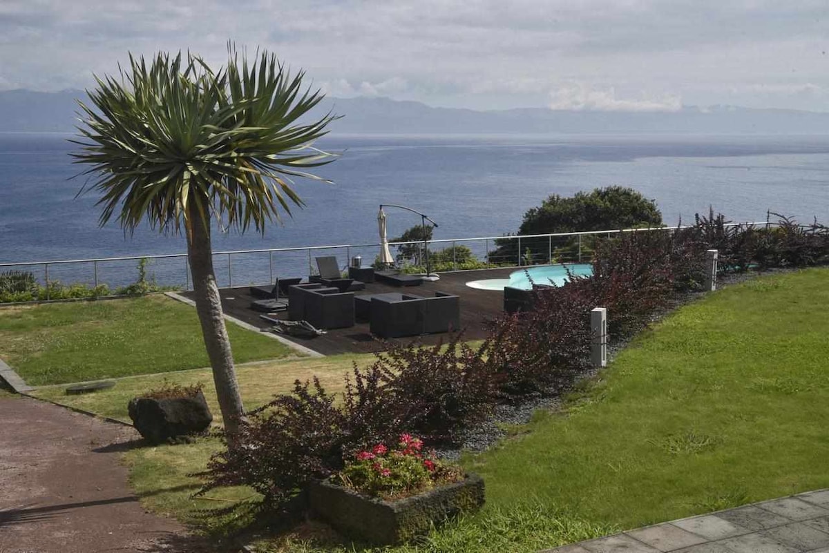 A well-maintained garden area offers a view of the expansive Atlantic Ocean. A palm tree is featured prominently, alongside modern lounge furniture surrounding a circular swimming pool. Lush greenery and flowering plants add to the serene atmosphere.