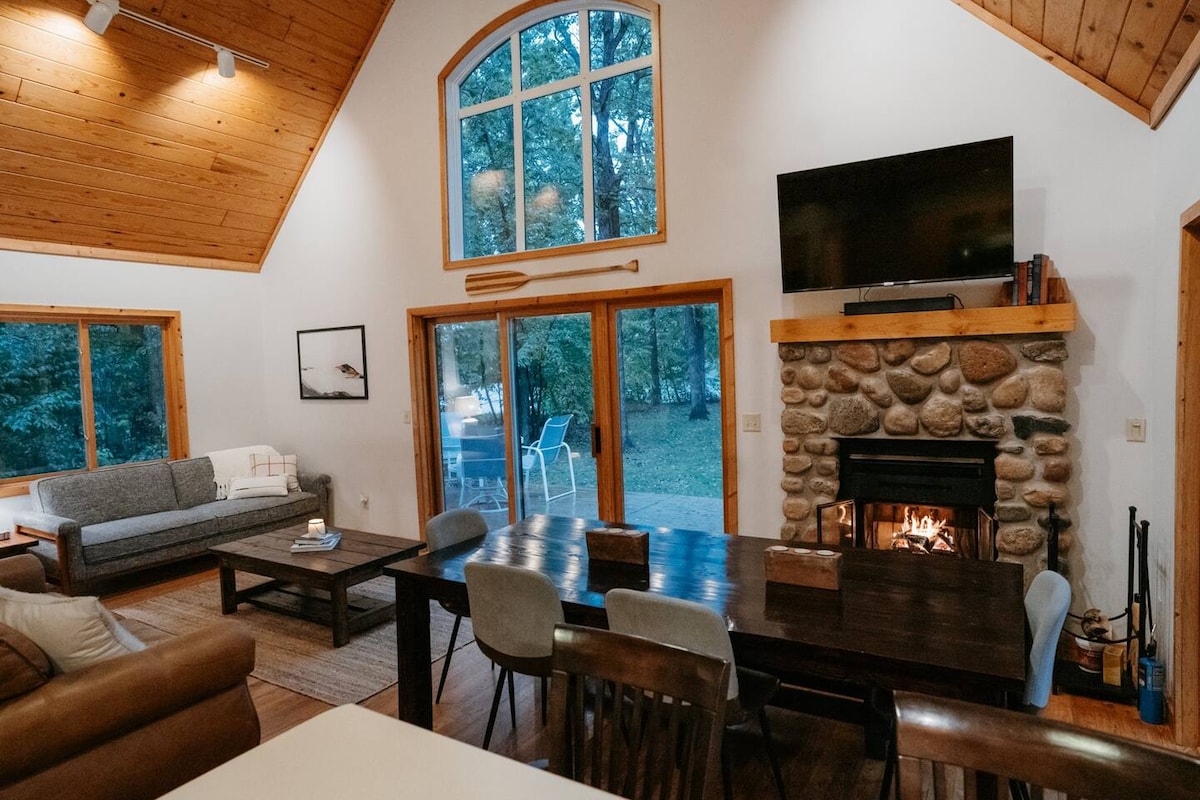 The living and dining area features vaulted wooden ceilings and large windows offering views of the surrounding trees. A stone fireplace with a wood-burning fire is set between wooden shelves, while a comfortable sofa and a dining table accommodate guests.