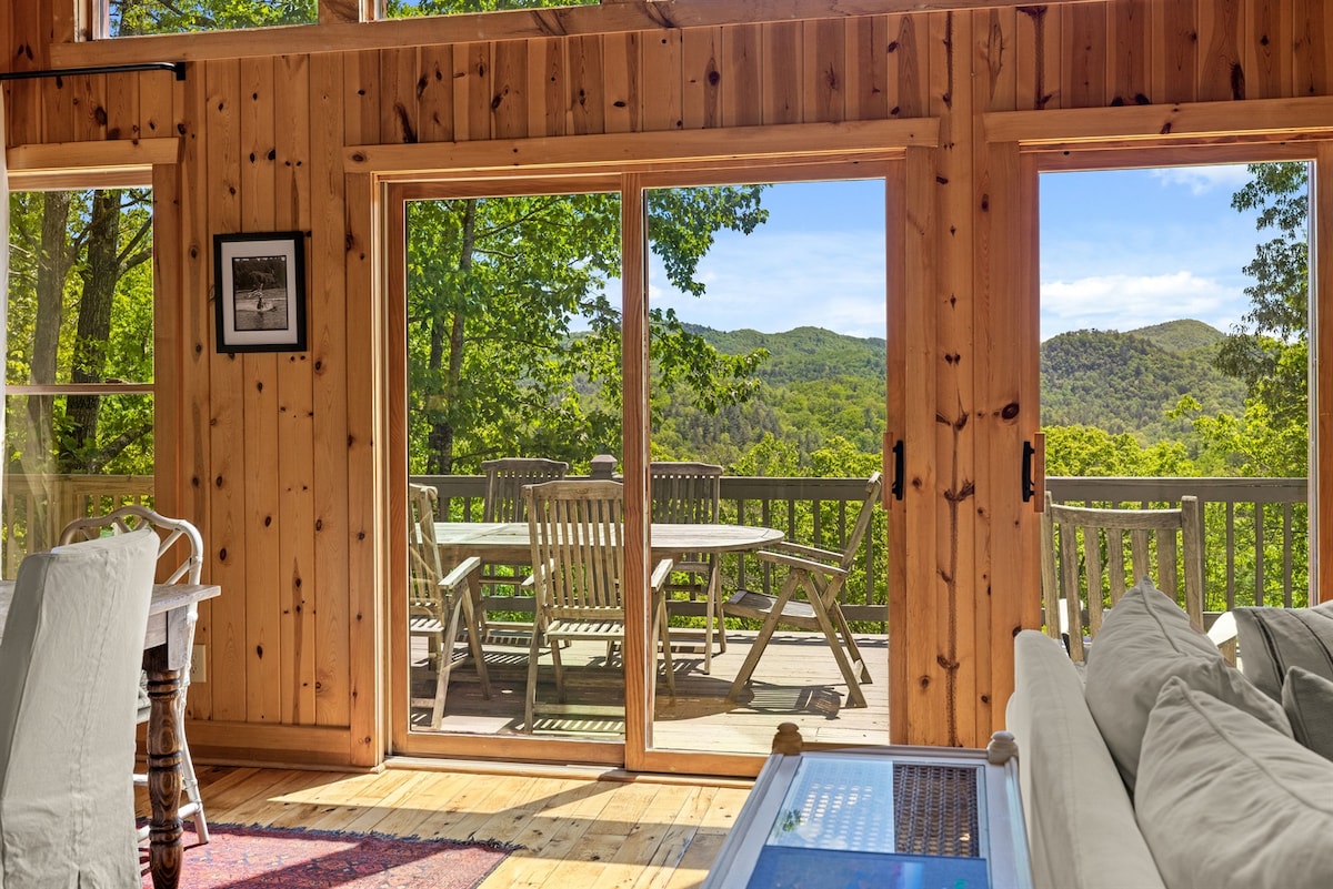Large sliding glass doors open to a spacious deck with outdoor seating and views of surrounding trees and hills. The interior features warm wood paneling and natural lighting, complemented by a cozy seating area with light-colored furniture.